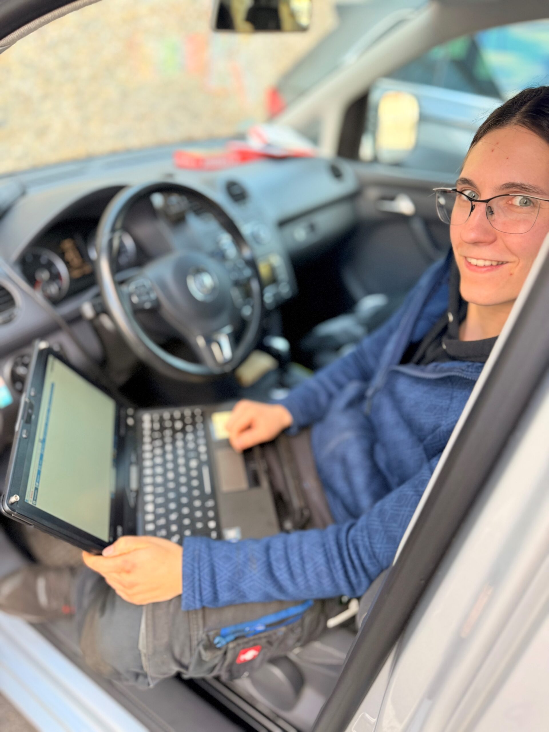Junge Frau mit Brille sitzt lächelnd auf dem Fahrersitz eines Autos und hält ein Laptop in der Hand. Sie trägt einen blauen Pullover. Im Hintergrund sind das Lenkrad, das Armaturenbrett und weitere unscharfe Fahrzeuginnenausstattung zu sehen.