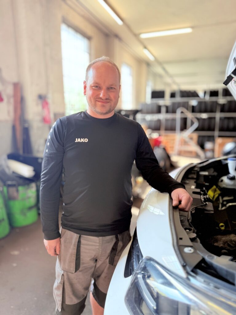 Mann mittleren Alters steht in einer Autowerkstatt und lehnt an der Motorhaube eines weißen Autos. Er trägt ein schwarzes Langarmshirt mit der Aufschrift "JAKO" und graue kurze Arbeitshose. Im Hintergrund sind unscharfe Werkstatteinrichtungen, Fenster mit Tageslicht und Regale mit Reifen zu sehen.