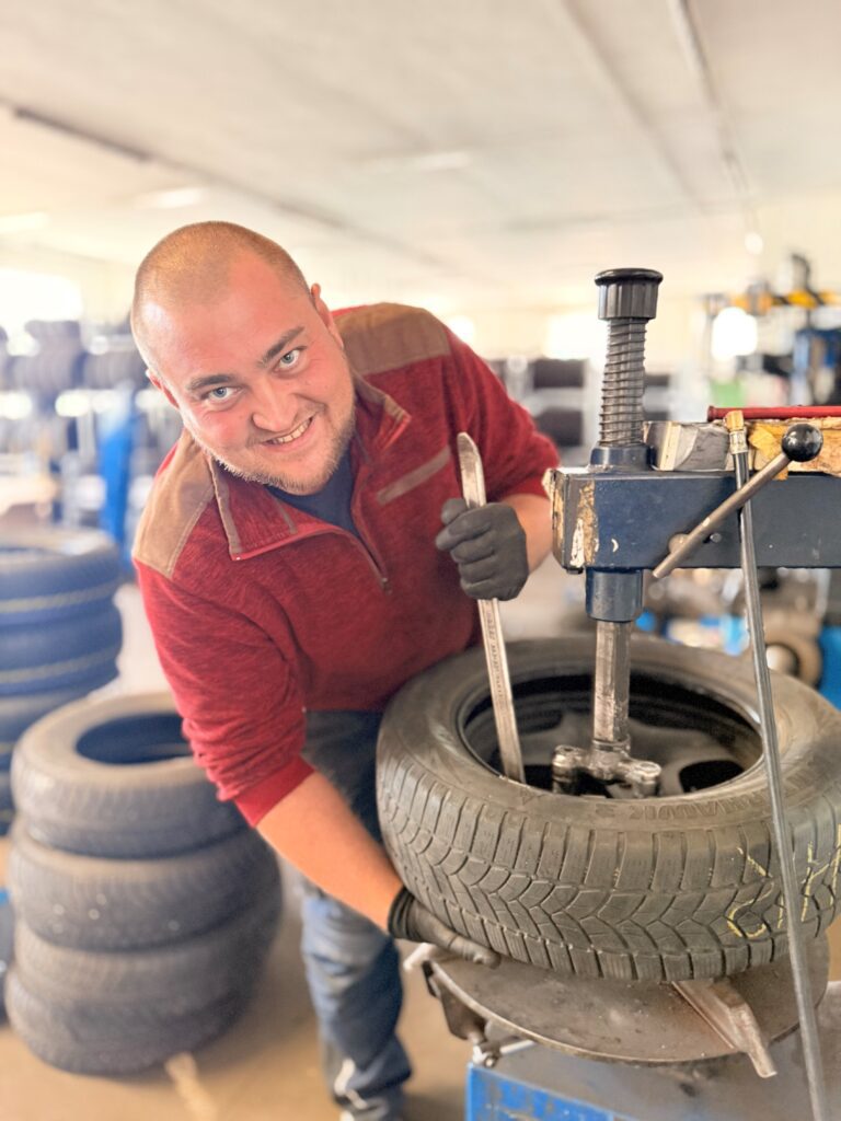 Mann mit Glatze und Bart lächelt in die Kamera, während er an einer Reifendemontiermaschine in einer Werkstatt arbeitet. Er trägt ein rotes Sweatshirt und schwarze Arbeitshandschuhe. Ein Auto-Reifen ist in der Maschine eingespannt. Im unscharfen Hintergrund sind weitere Reifen und Werkstatteinrichtungen zu sehen.