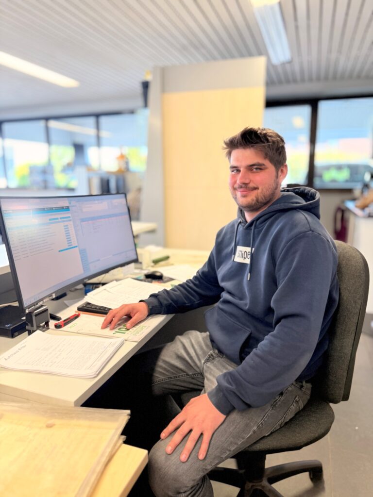 Junger Mann sitzt an einem Schreibtisch in einem Büro und schaut zur Kamera. Er trägt einen dunkelblauen Hoodie und graue Jeans. Vor ihm steht ein Monitor, daneben liegen Unterlagen. Im Hintergrund sind unscharf große Fenster mit Blick nach draußen sowie gelbe Wände zu sehen.
