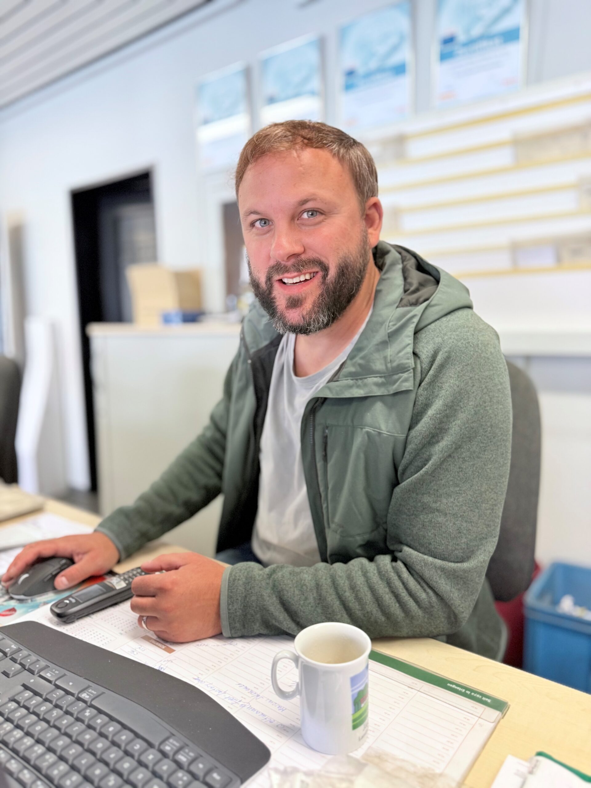 Mann mit Bart und braunen Haaren sitzt lächelnd an einem Schreibtisch in einem hellen Büro. Er trägt eine olivgrüne Jacke über einem grauen T-Shirt. Auf dem Schreibtisch stehen eine weiße Tasse und Unterlagen, daneben liegen eine Computermaus und ein Telefon. Im Hintergrund sind Fenster und unscharfe Büroeinrichtungen zu sehen.
