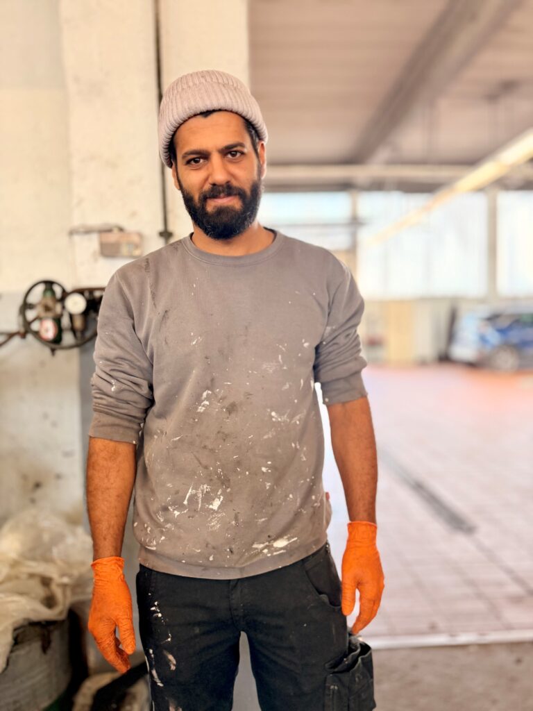 Mann mit Bart und Mütze steht in einer Werkstatt. Er trägt einen grauen Pullover mit Farbflecken, eine schwarze Hose mit weißen Flecken und orangefarbene Arbeitshandschuhe. Im unscharfen Hintergrund ist eine Werkstatt zu sehen.