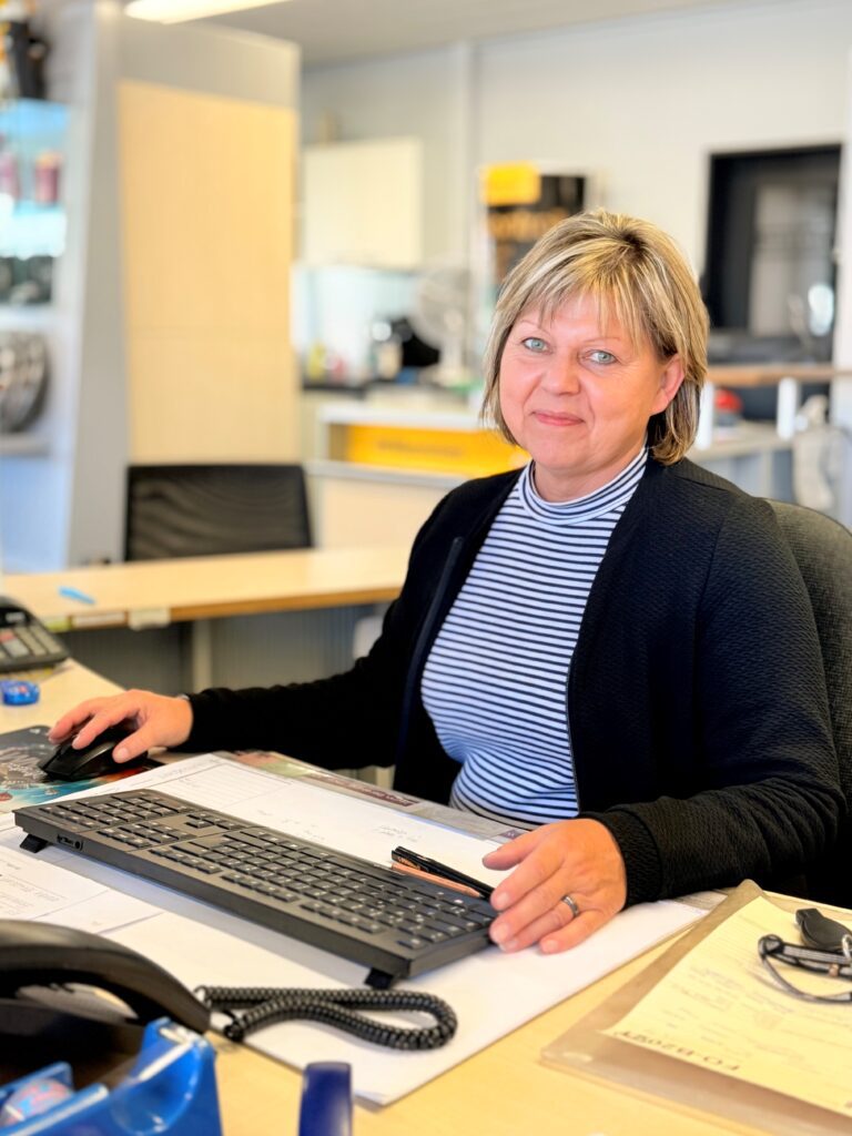 Frau mittleren Alters mit blondem Haar sitzt an ihrem Schreibtisch in einem Büro. Sie trägt einen dunklen Cardigan über einem blau-weiß gestreiften Shirt und hat ihre Hand auf einer Computermaus. Vor ihr liegen Dokumente und ein Stift. Im Hintergrund sind unscharfe Büroeinrichtungen sowie gelbe und weiße Schränke zu sehen.
